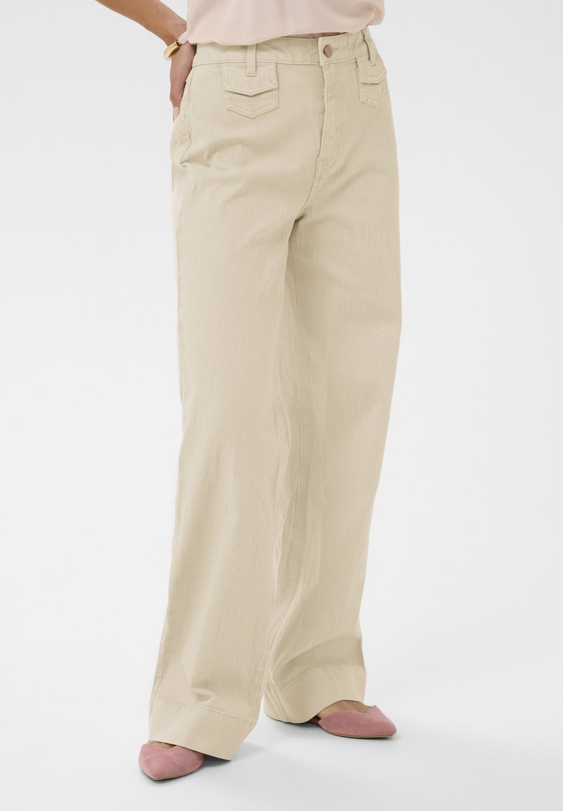 Beige wijde broek met zakken aan de voor- en achterkant, gedragen met roze slip-on schoenen met spitsen neus en een lichte blouse.