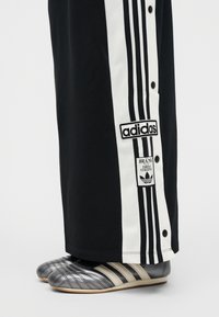 Πόδια που φορούν μαύρο παντελόνι Adidas με λευκές ρίγες και κουμπιά, συνδυασμένο με ασημί αθλητικά παπούτσια Adidas με τρεις λευκές ρίγες.