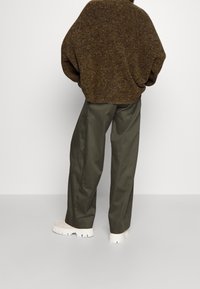 Übergroßer brauner, strukturierten Strickpullover kombiniert mit lockeren olivgrünen Hosen und weißen Schuhen mit dicker Sohle. Neutraler Hintergrund.