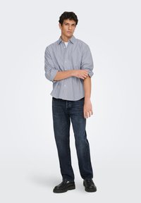 Ριγέ πουκάμισο με κουμπιά σε μπλε και άσπρο, συνδυασμένο με σκούρο τζιν τζιν. Το μοντέλο φοράει μαύρα παπούτσια και στέκεται με τα χέρια σταυρωμένα.