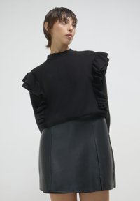 Pull noir avec des manches volantes, associé à une jupe en cuir noir ajustée. Les textures contrastent, mettant en valeur la structure et la silhouette.