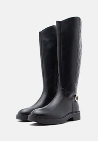 Pár černých kožených vysokých bot po kolena s plochou podrážkou, bočním zipem, prošívaným vzorovaným zadním panelem a dekorační sponou blízko paty.