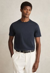 Mann, der ein marineblaues kurzärmeliges T-Shirt in eine weiße Hose mit schwarzem Gürtel gesteckt trägt, steht vor einem schlichten hellen Hintergrund.