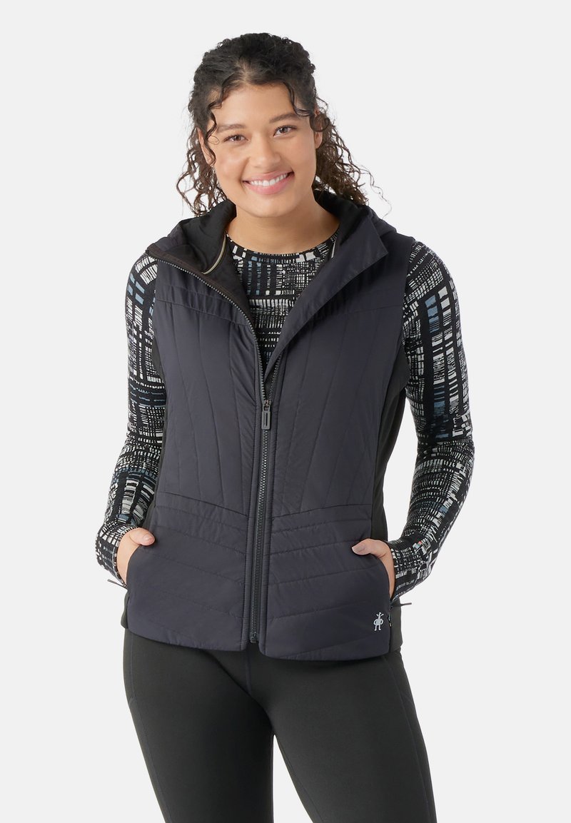 Black quilted vest with a hood, featuring zip closure and two side pockets. Worn over a patterned long-sleeve top. Smooth texture.