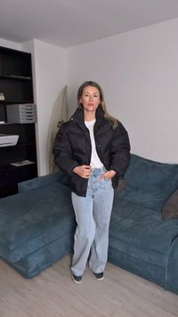 Black puffer jacket with high collar and snap buttons, paired with light blue jeans. The background features a green couch and shelving.
