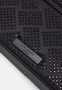 Sac à main noir avec une surface texturée ornée de clous noirs regroupés et une plaque en métal rectangulaire portant le logo "Kurt Geiger London."