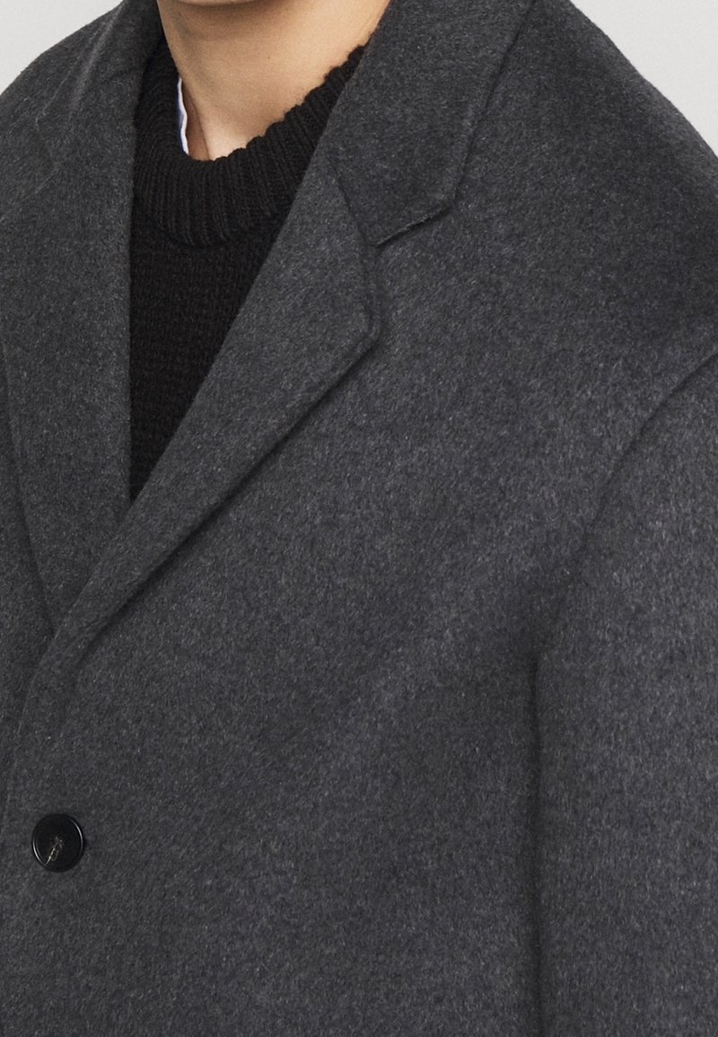 Manteau en laine gris foncé à larges revers porté sur un pull noir en maille à encolure ronde.