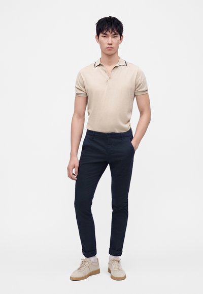 Homme debout, une main dans la poche, portant un polo beige à manches courtes, un pantalon slim foncé, des chaussettes blanches et des baskets beiges sur fond blanc.