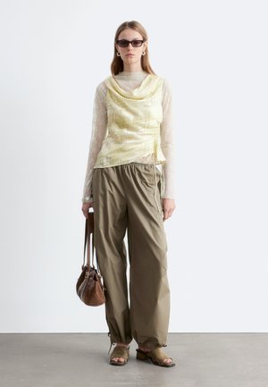 Žena má na sebe priehľadný top s dlhým rukávom pod žltou vzorovanou blúzkou bez rukávov, voľné hnedé nohavice, zelené sandále s otvorenou špičkou, drží hnedú kabelku.