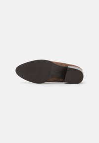 Zapato de ante marrón con suela de goma. Presenta una punta redondeada y una superficie texturizada. El tacón es apilado para mayor estabilidad.