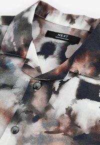 Camisa de manga corta con un patrón abstracto en tonos de negro, marrón y blanco. Tiene un cuello y dos botones oscuros. La etiqueta dice "NEXT."