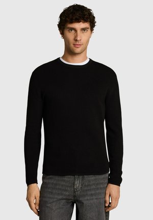 Mann mit kurzen lockigen Haaren, der einen schwarzen Langarmpullover über einem weißen Unterhemd und graue Jeans trägt, stehend vor einem einfarbigen Hintergrund.