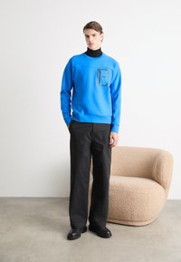 Giovane uomo in piedi accanto a una poltrona beige testurizzata, indossa un sudario blu sopra un dolcevita nero, pantaloni neri e scarpe nere in una stanza minimalista.