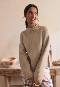 Beige bordázott kötött pulóver magas nyakkal és hosszú, széles ujjal, kockás alsók felett viselve. Sima textúrájú, laza fazonú, minimális dizájnnal.