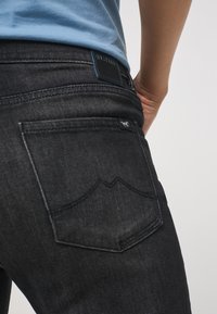 Mörk denimjeans har en bakficka med en vinkelaktig design, stickade detaljer och en läderetikett i midjan. Tyget har en slät textur.