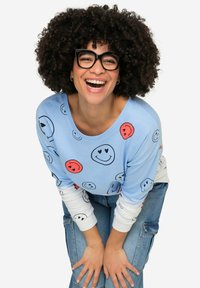 Camisa de manga larga azul claro con estampados de caras sonrientes, superpuesta a una camiseta blanca. Lleva puestos unos vaqueros de mezclilla azul. Cabello rizado y gafas negras.