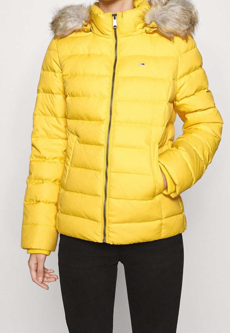 Żółta pikowana kurtka z suwakiem z przodu, wykończeniem kaptura z sztucznego futra, elastycznymi mankietami i bocznymi kieszeniami, o teksturowanej powierzchni i wzorze pikowanym.