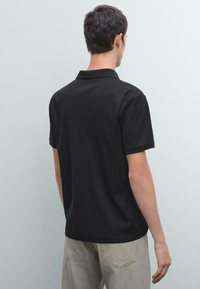 Sort bomulds polo skjorte med korte ærmer og krave, vist fra ryggen, parret med lyse bukser. Enkel design og tekstur.