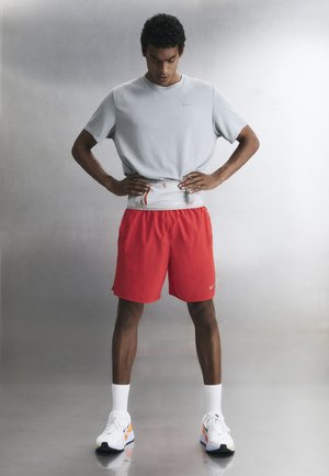 Mann i røde shorts og grå skjorte justerer en grå midjeveske, står på et glatt gulv mot en nøytral bakgrunn.