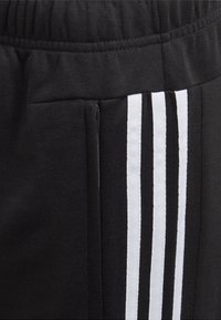 Los pantalones de chándal negros cuentan con una cinturilla texturizada, un bolsillo con cremallera y tres rayas blancas verticales a lo largo del costado.