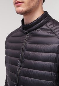 Svart vadderad jacka med hög krage, frontdragkedja och ribbad textur med horisontella sömmar. Lätt material.