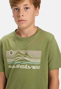 Alyvuogių žalios spalvos medvilnės marškinėliai su grafikų piešiniu, kuriame pavaizduoti kalnai, saulė ir bangos pastelinių spalvų. Po piešiniu yra "Quiksilver" logotipas.