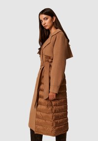 Hnedý dvojitý kabát s vrchnou časťou z vlny a dolnou časťou vyplnenou páperím, s opaskom, širokými klopami a prešívanou textúrou.