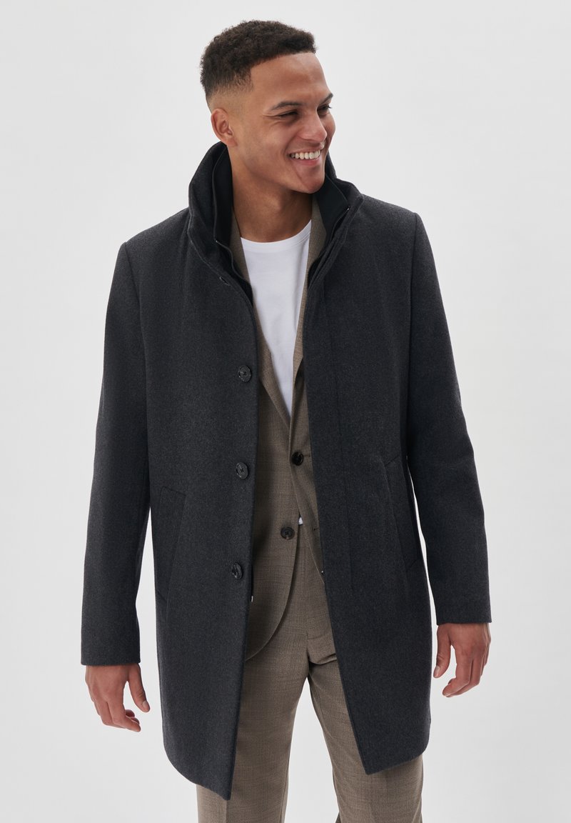 Abrigo de lana gris oscuro con cuello alto, botones delanteros y bolsillos laterales, llevado sobre un traje beige y una camisa blanca. Textura suave.