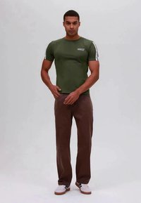 Grünes kurzärmliges T-Shirt mit seitlichen Streifen, kombiniert mit braunen Hosen und weißen Sneakern mit blauen und schwarzen Akzenten.