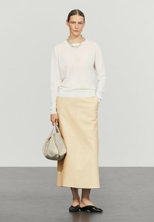 Vrouw draagt witte top met lange mouwen, beige midirok, zwarte platte schoenen, zilveren ketting, houdt beige handtas vast met hand in zak.