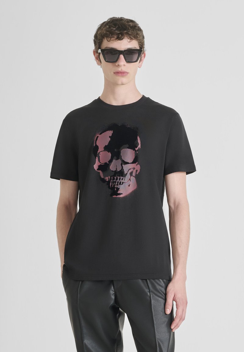 Camiseta negra de manga corta con un gráfico central de calavera en tonos de rosa y negro. Se lleva con pantalones negros brillantes.