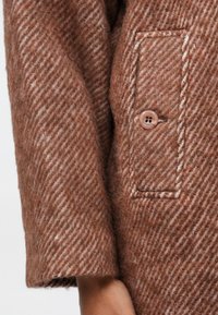 Manga de um casaco de lã em padrão espinha de peixe castanho e branco, com um botão castanho de quatro furos e uma aba de bolso retangular costurada.