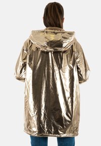 Metalen gouden hoodie-jack met een glanzende afwerking, voorzien van een pluche gevoerde capuchon, een losse pasvorm en een lengte tot onder de heupen. Gestructureerd oppervlak is zichtbaar.
