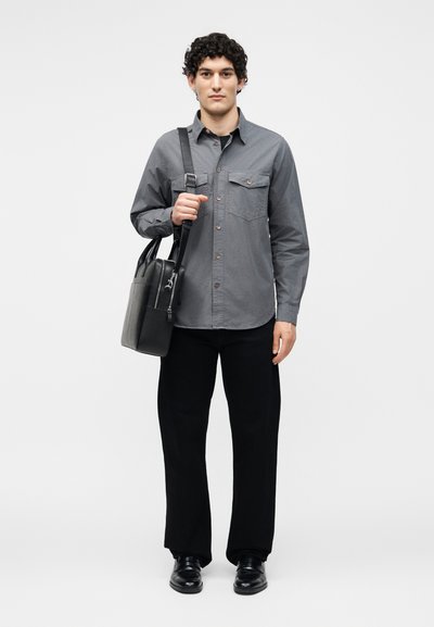 Νέος άνδρας με σγουρά μαλλιά, ντυμένος με γκρι πουκάμισο με κουμπιά, μαύρο παντελόνι, μαύρα παπούτσια και κρατώντας μια μαύρη και γκρι τσάντα ώμου.
