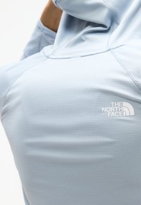 Άτομο που φοράει ανοιχτό μπλε φούτερ The North Face, φαίνεται από πίσω και σηκώνει το χέρι προς το κεφάλι.