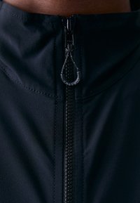 Svart sykkeljakke med høy krage, med hel lengde glidelås og strukturert stoff. Glidelåsens drag har "Castelli" -merket.