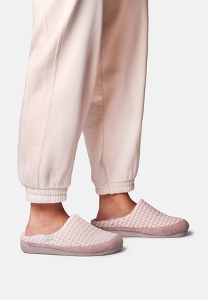 Personne portant des chaussons légers rose clair texturés et un pantalon rose pâle avec revers, debout sur un fond uni.