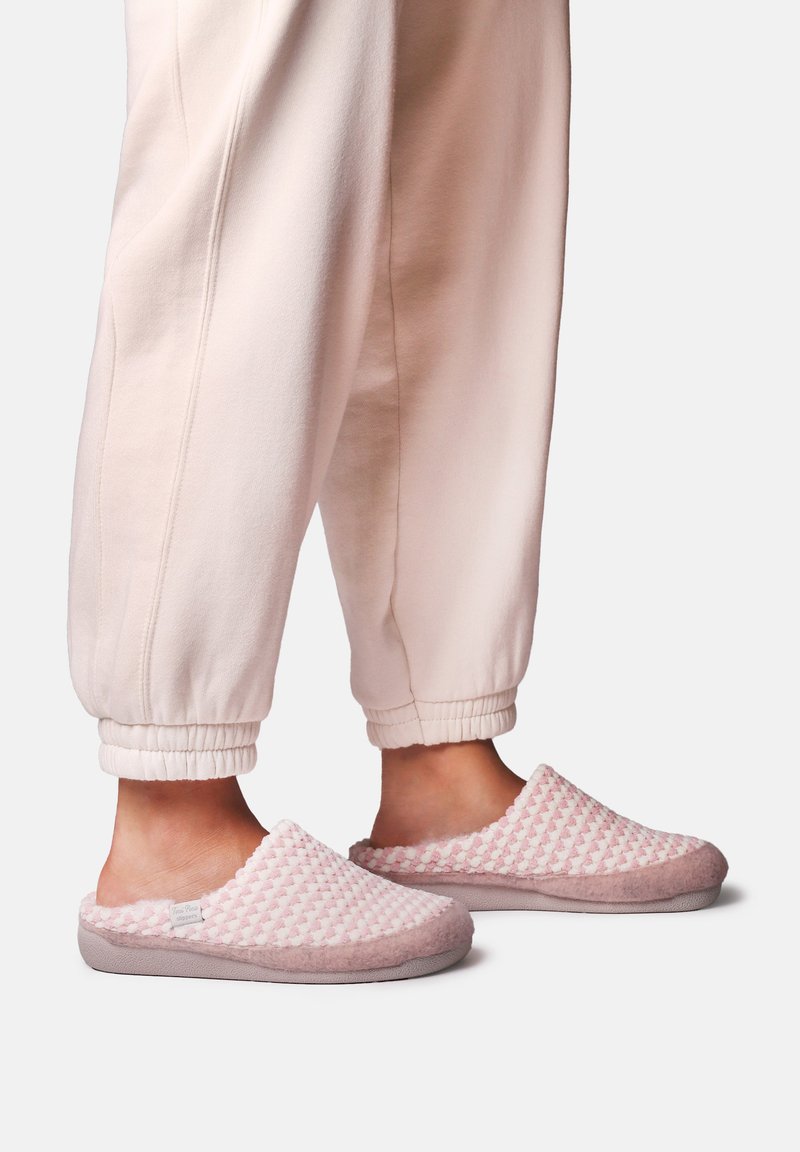 Personne portant des chaussons légers rose clair texturés et un pantalon rose pâle avec revers, debout sur un fond uni.