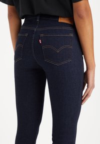 Jeans de mezclilla azul oscuro con cintura alta, que cuentan con dos bolsillos traseros y costuras naranjas contrastantes. Se puede ver la etiqueta de la marca de cuero.