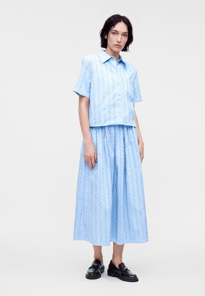 Femme portant une chemise bleu clair à manches courtes boutonnée et une jupe midi assortie avec de petits motifs ajourés, associée à des mocassins noirs.