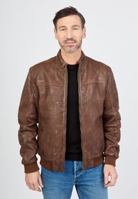Bruine leren bomberjack met ribgebreide manchetten en zoom, een ritssluiting aan de voorkant en een licht gestructureerde afwerking. Draagt over een zwart shirt.