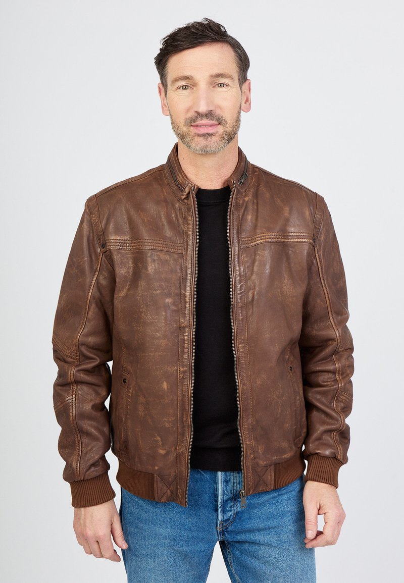 Bruine leren bomberjack met ribgebreide manchetten en zoom, een ritssluiting aan de voorkant en een licht gestructureerde afwerking. Draagt over een zwart shirt.