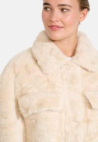 Bežna jakna iz umetnega krzna z mehko teksturo, ki ima velik ovratnik in dve sprednji žepni, ter prikazuje kvadratno zasnovo.