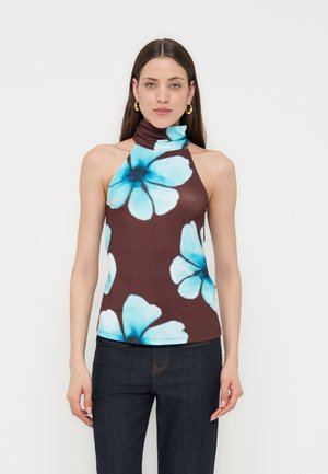 Młoda kobieta ubrana w brązowy, bezrękawnikowy top z dużymi niebieskimi motywami kwiatowymi, połączony z ciemnoniebieskimi dżinsami, stoi na tle jednolitego tła.