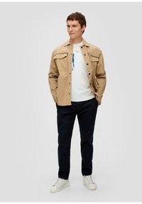 Beżowa koszula z guzikami i kieszeniami na klapki, noszona na białym t-shircie z grafiką, w połączeniu z granatowymi spodniami i białymi sneakersami, stojąca na neutralnym tle.