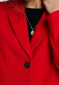 Frau mit einem roten Mantel mit schwarzem Knopf über einem schwarzen Oberteil, die eine silberne Halskette mit einem runden Anhänger trägt.