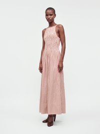Woman wearing sleeveless pink and white checkered maxi dress with smocked bodice and brown high-heeled shoes standing on a plain background.