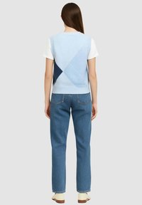 Maglione senza maniche blu chiaro con motivi geometrici, indossato sopra una camicia bianca, abbinato a jeans blu a gamba dritta e scarpe beige.