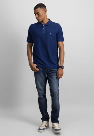 Mężczyzna w granatowej koszulce polo, ciemnoniebieskich dżinsach, beżowych trampkach i czarnym zegarku, stojący z jedną ręką w kieszeni, patrzący w dół.