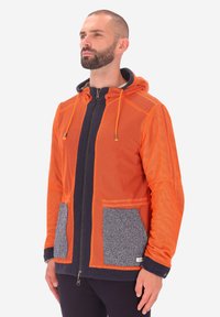 Uomo con barba che indossa una giacca zip-up arancione in rete con tasche in maglia grigia e rifiniture blu navy, in piedi e che guarda leggermente in alto e a destra.